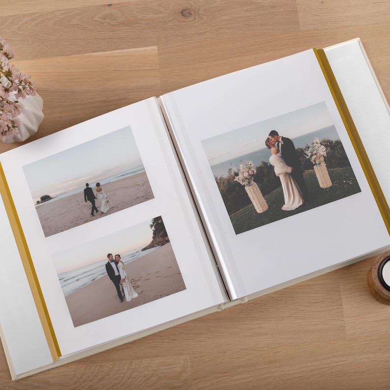 May include: An open photo album with white pages and gold spine accents, displaying three wedding photographs. The images show a couple in formal attire on a beach and at an outdoor ceremony. The album is laid flat, showcasing the printed photos.