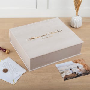 May include: A light beige, rectangular keepsake box with the names "Allison and Matthew" and the date "July 12, 2023" in gold lettering. A photo, a card, and a wax seal are on the wooden surface.
