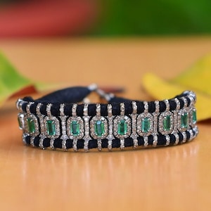 May include: A black bracelet with emerald-cut green gemstones set in a silver-colored metal. The bracelet has a woven black band and is displayed on a light brown surface.