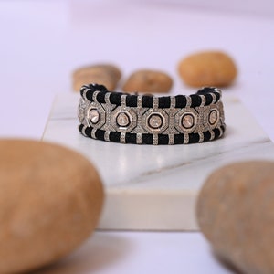 May include: A black and silver bracelet with a geometric design. The bracelet features a black woven band and silver-colored geometric shapes with clear stones. The bracelet is displayed on a white surface with brown stones in the background.