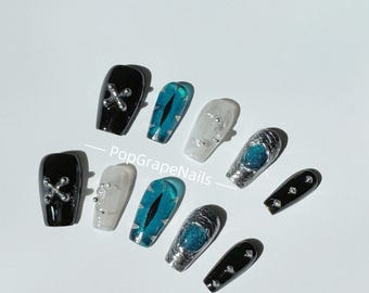 May include: A collection of press-on nails in various designs. Some nails are black with silver cross accents, others feature a teal eye design, and some are clear with silver embellishments. The nails are coffin-shaped.