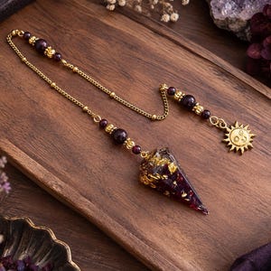 Può includere: Un pendolo con una punta conica in resina viola scuro con scaglie di foglia d'oro, attaccato a una catena color oro. La catena è decorata con perline viola scuro e un ciondolo a forma di sole color oro all'estremità, un oggetto decorativo.