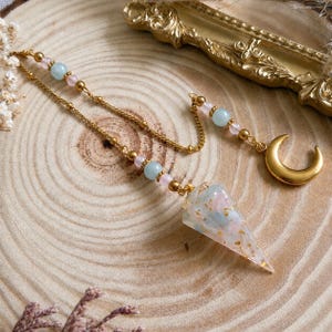 May include: A pendulum featuring a conical resin point with embedded gold flakes, light blue and pink beads, and a gold-toned chain. The chain includes a small gold-toned crescent moon charm and additional light blue and pink spherical beads.