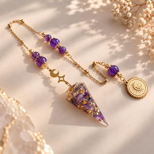 May include: A pendulum featuring a cone-shaped pendant made of purple amethyst and gold flakes, suspended from a gold chain. The chain includes round purple amethyst beads, a small gold crescent moon and star charm, and a round gold sun medallion.