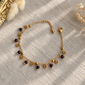 Peut inclure: Un bracelet à chaîne doré orné de perles sphériques rouge foncé alternant avec de petites breloques en forme de fleurs dorées. Le bracelet est doté d'un fermoir mousqueton et d'une section de chaîne réglable à une extrémité.