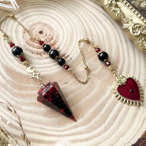 May include: A pendulum with a red and black crystal point, gold chain, and black bead accents. The pendulum features a gold crescent moon and star charm. A heart-shaped charm with a red centre and gold sunburst design is also present.