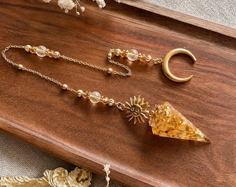 May include: A gold-toned pendulum featuring a conical resin point filled with gold flakes, attached to a chain with clear beads and a sun charm. The chain ends in a gold-toned crescent moon. The design incorporates metallic gold accents and clear spherical beads throughout the length of the chain, creating a cohesive piece of jewelry with celestial motifs.