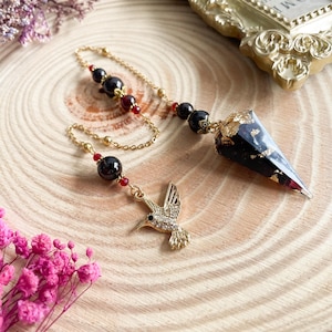 Può includere: Un pendolo nero e oro con un ciondolo a forma di colibrì. Il pendolo è realizzato in resina e ha una finitura in foglia d'oro. La catena è in metallo dorato e ha perline rosse.