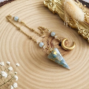 May include: A pendulum featuring a cone-shaped pendant made of blue and white marbled resin with gold leaf flakes. The pendant hangs from a gold-toned metal chain accented with light blue spherical beads, clear crystals, and a gold-toned crescent moon charm.