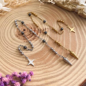 May include: Three necklaces with beaded chains and star or cone pendants. One necklace is silver-toned with gray beads, another is gold-toned with gold beads, and the third is gold-toned with gray beads. The necklaces are displayed on a wooden surface.