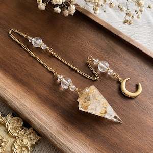 May include: A pendulum featuring a clear, cone-shaped resin point with gold-coloured flakes inside, suspended from a gold-toned metal chain. The chain includes clear spherical beads and terminates in a small, polished gold-coloured crescent moon charm.