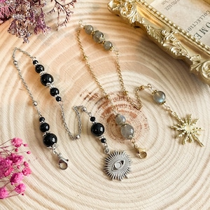 May include: Three silver chains with charms and beads. The chains feature black beads, grey beads, and a silver eye charm. The chains are displayed on a wooden surface.