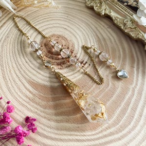 May include: A gold-colored, ornate pendant with a clear, teardrop-shaped resin body embedded with gold leaf. The pendant is attached to a delicate gold chain adorned with clear and pink beads, and a small heart-shaped charm.