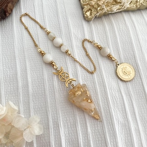 Può includere: Un pendolo color oro con un cristallo trasparente e appuntito e scaglie d'oro. Il pendolo presenta una catena dorata con perline bianche e un ciondolo dorato con un design a tripla luna e stella. È incluso anche un ciondolo a moneta d'oro con un disegno del sole.