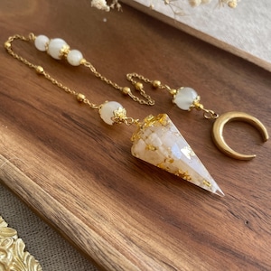 Può includere: Un pendolo color oro con un cristallo a forma di cono con macchie bianche e dorate. Il pendolo ha una catena con perline bianche e un ciondolo a mezzaluna. Il design è su uno sfondo di legno.