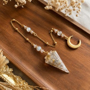 May include: A pendulum featuring a clear, cone-shaped resin point filled with gold flakes, attached to a gold-toned chain. The chain is adorned with small, iridescent round beads and clear crystals, ending in a polished gold-toned crescent moon charm.