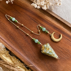 Può includere: Un pendolo con un ciondolo conico composto da frammenti di pietra verde chiaro e scaglie dorate, fissato a una catena color oro. La catena è decorata con piccole perline verdi e dettagli dorati, terminando con un ciondolo a mezzaluna color oro.