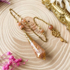 Op de afbeelding: Een pendel met een perzikkleurige, gefacetteerde kristallen punt met bladgoudvlokken, bekroond met een goudkleurige metalen dop. De ketting bevat perzikkleurige kralen en eindigt in een goudkleurige metalen sleutelbedel met een hartvorm.
