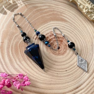 Può includere: Un pendolo divinatorio con una punta di cristallo blu scuro e una catena d'argento. La catena è decorata con perline nere e blu. Un ciondolo d'argento con un disegno di sole e luna è attaccato alla fine della catena.