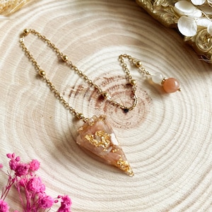 Op de afbeelding: Een gouden kettingpendulum met een perzikkleurige kristallen punt en gouden vlokken. De ketting heeft kleine gouden kralen en een perzikkleurige kristallen kraal aan het einde.