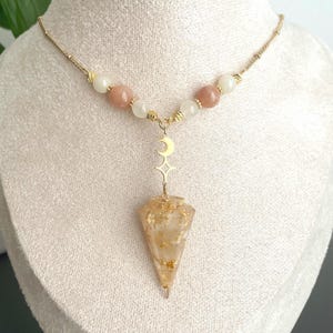 Puede incluir: Collar de oro con un dije de oro en forma de media luna y estrella, y un dije de cristal de cuarzo transparente con destellos dorados. El collar está hecho con cuentas blancas y melocotón.