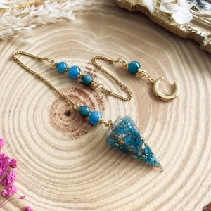Op de afbeelding: Een pendel met een gouden ketting en een turquoise, kegelvormige kristallen hanger. De ketting is versierd met blauwe kralen. Een gouden halve maan bedel hangt aan het einde. De hanger heeft gouden vlokken.