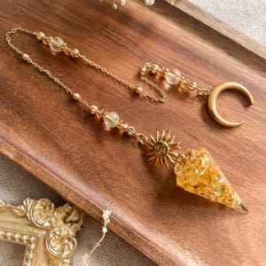 以下が含まれることがあります： 三日月、太陽、金箔入りのクリスタルコーンが付いたゴールドカラーのサンキャッチャー。太陽は笑顔です。サンキャッチャーは、透明なクリスタルビーズが付いた繊細なゴールドチェーンから吊り下げられています。木製のトレイに飾られています。