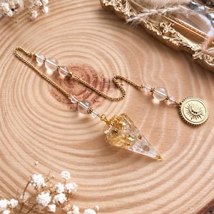 Könnte beinhalten: Ein Pendel mit einer klaren, kegelförmigen Harzspitze, die mit Goldflocken gefüllt ist und an einer goldfarbenen Kette hängt. Die Kette ist mit klaren, runden Perlen verziert und endet in einem kleinen, runden goldfarbenen Anhänger mit Sonnen- und Mondmotiv.