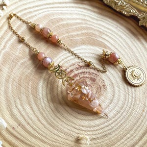 Peut inclure: Un pendule doré avec une chaîne et des perles couleur pêche. Le pendule présente un cristal clair et pointu avec des paillettes dorées et un charme de triple lune. Une pièce d'or avec un motif de soleil et de lune est suspendue au bout de la chaîne. Le pendule repose sur une surface en bois.