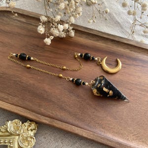 Könnte beinhalten: Ein Pendel mit einem schwarzen, kegelförmigen Kristall, verziert mit Goldflocken, hängt an einer goldenen Kette. Die Kette ist mit einem goldenen Halbmond-Anhänger und schwarzen Perlen verbunden. Das Pendel liegt auf einem Holztablett.