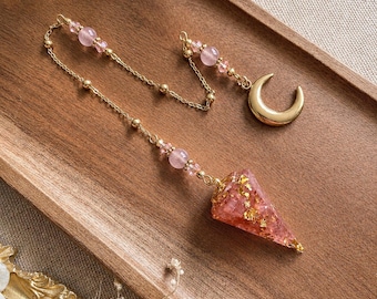 May include: A pendulum featuring a cone-shaped resin point infused with gold flakes, attached to a gold-toned chain. The chain is accented with light pink beads and a small gold-toned crescent moon charm at the end of the chain segment.