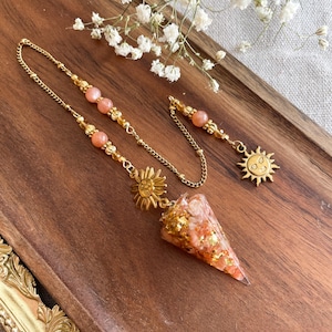 Könnte beinhalten: Ein Pendel mit einer kegelförmigen Harzspitze, die mit orangefarbenen und goldenen Flocken gefüllt ist, befestigt an einer goldfarbenen Kette. Die Kette enthält runde pfirsichfarbene Perlen, kleine goldene Zwischenstücke und einen Sonnenanhänger.
