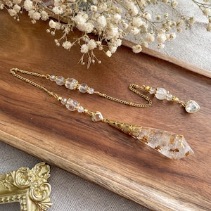 Può includere: Un pendolo color oro con una punta di cristallo trasparente e scaglie d'oro. Il pendolo è attaccato a una catena con perline di cristallo trasparente e un ciondolo a forma di cuore. L'articolo è esposto su un vassoio di legno con fiori bianchi sullo sfondo.