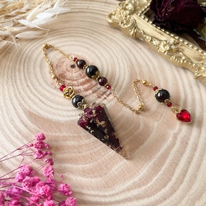 Könnte beinhalten: Ein Pendel mit einer goldenen Kette und einer roten und schwarzen Kristallspitze, die mit Blattgold verziert ist. Die Kette ist mit schwarzen und roten Perlen geschmückt.