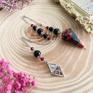 May include: A black and red pendulum with a silver chain and red and black beads. The pendulum is shaped like a teardrop and has a silver charm with a sun and moon design.