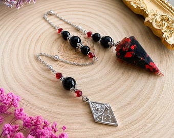 May include: A pendulum featuring a conical black and red resin point attached to a silver-toned chain. The chain includes black spherical beads, small red crystal accents, and a diamond-shaped silver charm engraved with a sun and moon design.