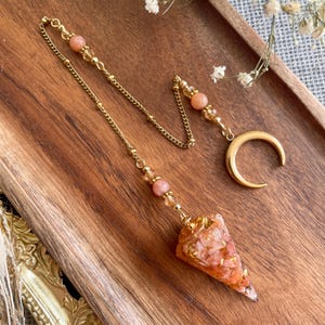 Peut inclure: Un pendule à chaîne en métal doré avec un pendentif conique rempli de éclats de pierre orange et rose et de flocons dorés. La chaîne est ornée de perles rondes couleur pêche et se termine par une breloque en forme de croissant de lune doré.