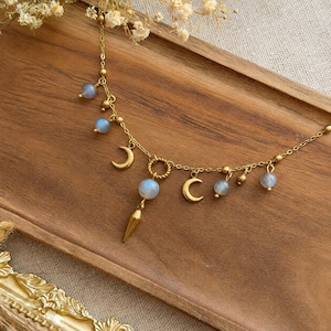 May include: A gold-toned necklace featuring a delicate chain adorned with small round blue-grey beads and several crescent moon charms. A central pendant consists of a larger round bead suspended above a pointed gold-toned metal spike charm.