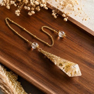 Könnte beinhalten: Ein Pendel mit einer klaren, facettierten Harzspitze, die mit Blattgoldflocken gefüllt ist und von einer goldfarbenen Filigrankappe gekrönt wird. Das Stück ist an einer goldfarbenen Kette mit zwei klaren, kugelförmigen Perlen befestigt.