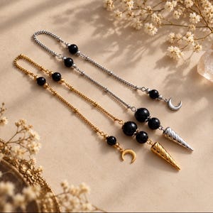Könnte beinhalten: Zwei Metallketten mit schwarzen, kugelförmigen Perlen, eine in Gold- und eine in Silberoptik. Jede Kette endet in einem spitzen, konischen Anhänger und einem Mondsichel-Charm, wobei weitere schwarze Perlen entlang der Ketten eingearbeitet sind.