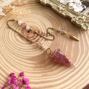 Op de afbeelding: Een goudkleurige pendel met een roze, kegelvormige kristallen hanger. De ketting heeft roze kralen en een puntig uiteinde. De pendel wordt gepresenteerd op een houten oppervlak, met een decoratief gouden frame op de achtergrond.
