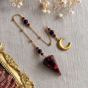 May include: A gold-toned chain pendulum featuring a pointed, dark resin pendant with embedded red flecks. The chain is accented with small black and red beads and terminates in a smooth, gold-toned crescent moon charm at the opposite end.