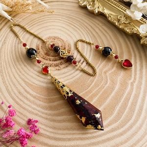 Peut inclure: Un pendule composé d'une pointe facettée en forme de cône rouge foncé avec des éclats de feuilles d'or, surmontée d'un capuchon en filigrane doré. La chaîne dorée comporte des perles noires, des perles rouges, une rose et un cœur rouge.