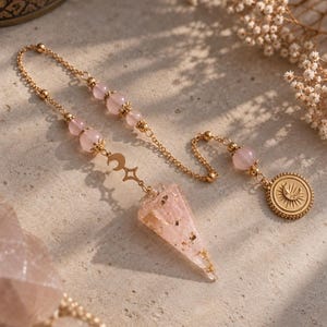 Op de afbeelding: Een goudkleurige kettingpendel met een puntige rozenkwarts kristallen hanger met goudvlokken. De ketting is versierd met ronde rozenkwarts kralen, een kleine maansikkelbedel en een ronde medaillon met een reliëf van een zon en maan.