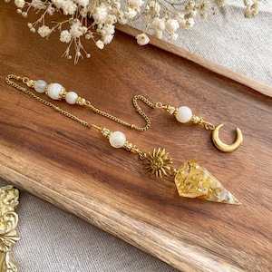Peut inclure: Un pendule doré avec un soleil et un croissant de lune, des perles blanches et un pendentif en résine transparent et pointu avec des paillettes dorées. Le pendule est suspendu à une chaîne.