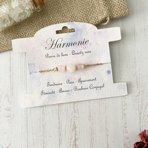 Peut inclure: Un bracelet en chaîne dorée avec trois perles roses et blanches. La carte indique "Harmonie", "Pierre de lune - Quartz rose", "Tendresse - Paix - Apaisement", et "Féminité - Amour - Bonheur Conjugal".