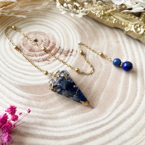 Könnte beinhalten: Goldfarbene Halskette mit blau-goldfarbenem Anhänger. Der Anhänger ist dreieckig und mit blauen Steinen und Goldflocken gefüllt. Am Ende der Kette befinden sich zwei blaue, kugelförmige Perlen.