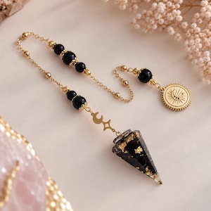 Könnte beinhalten: Ein Pendel bestehend aus einem schwarzen Harzkegel mit Goldflocken, aufgehängt an einer goldfarbenen Kette. Die Kette enthält schwarze kugelförmige Perlen, einen kleinen Mond- und Sternanhänger sowie einen runden goldfarbenen Anhänger mit Sonnen- und Mondmotiv.