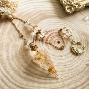 Puede incluir: Un péndulo de cristal transparente y dorado con una punta puntiaguda y una cadena con cuentas de cristal transparente. El péndulo cuelga de una cadena dorada con un dije de media luna.