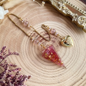 May include: A gold-colored chain with a pink and gold pendulum. The pendulum is a pink, triangular shape with gold flakes inside. A gold heart-shaped locket hangs from the chain. The chain has pink beads.